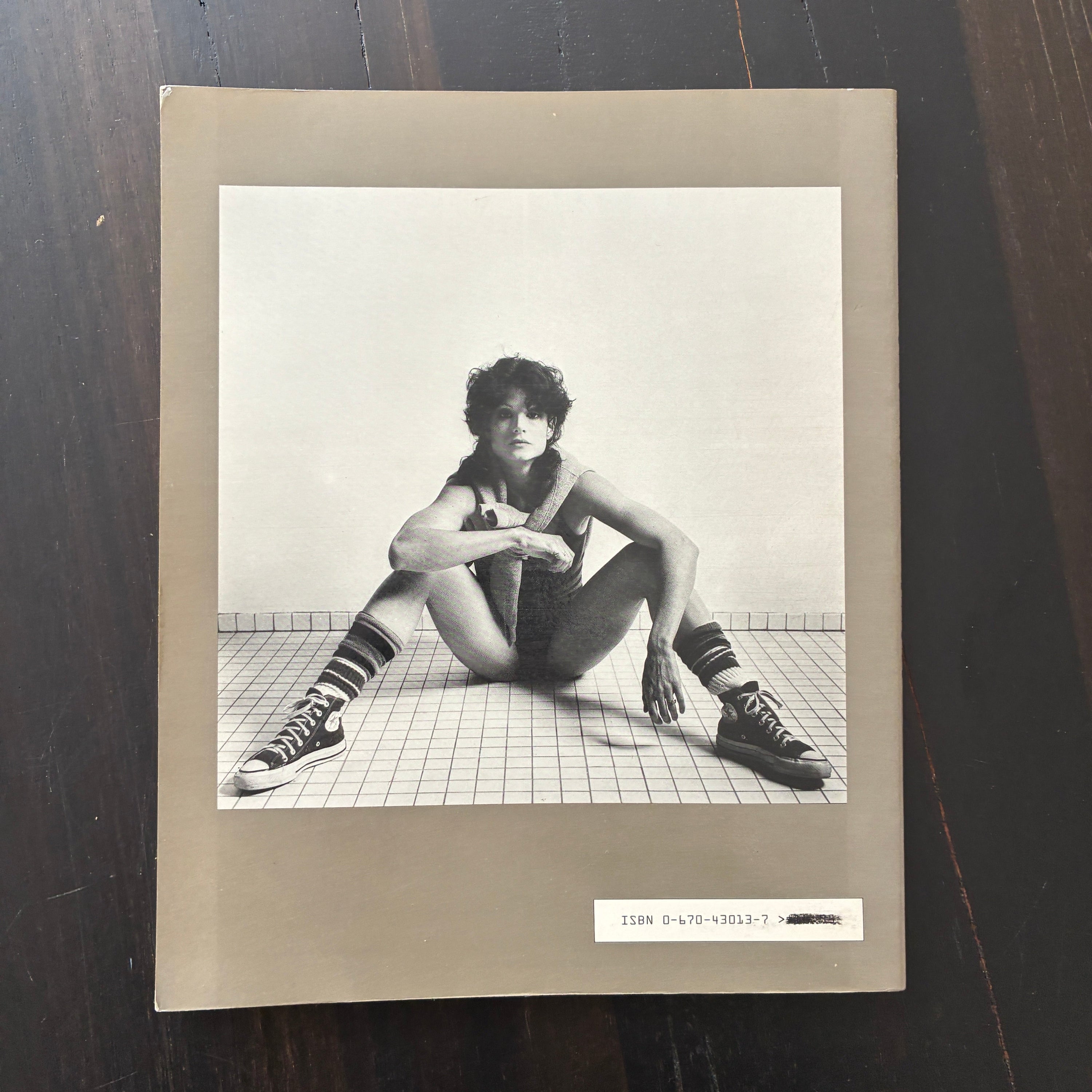 A square art print or album cover photographed from above on a dark wooden surface. The cover has a beige border and features a black-and-white photograph of a seated person on a tiled floor.