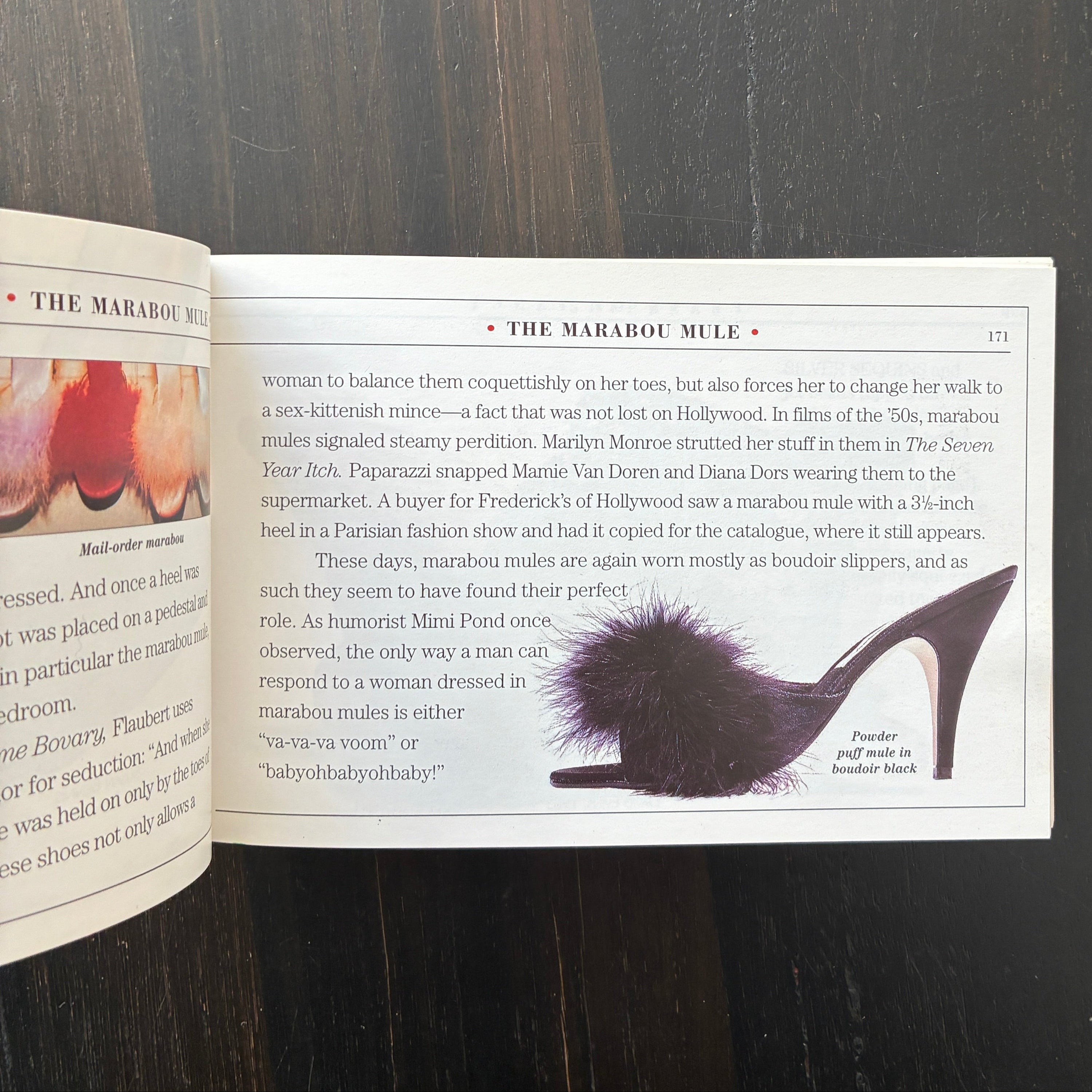 An open book lying on a dark wooden table, showing a page with text and a black-and-white fashion illustration of a high-heeled shoe with a large fluffy pom-pom.