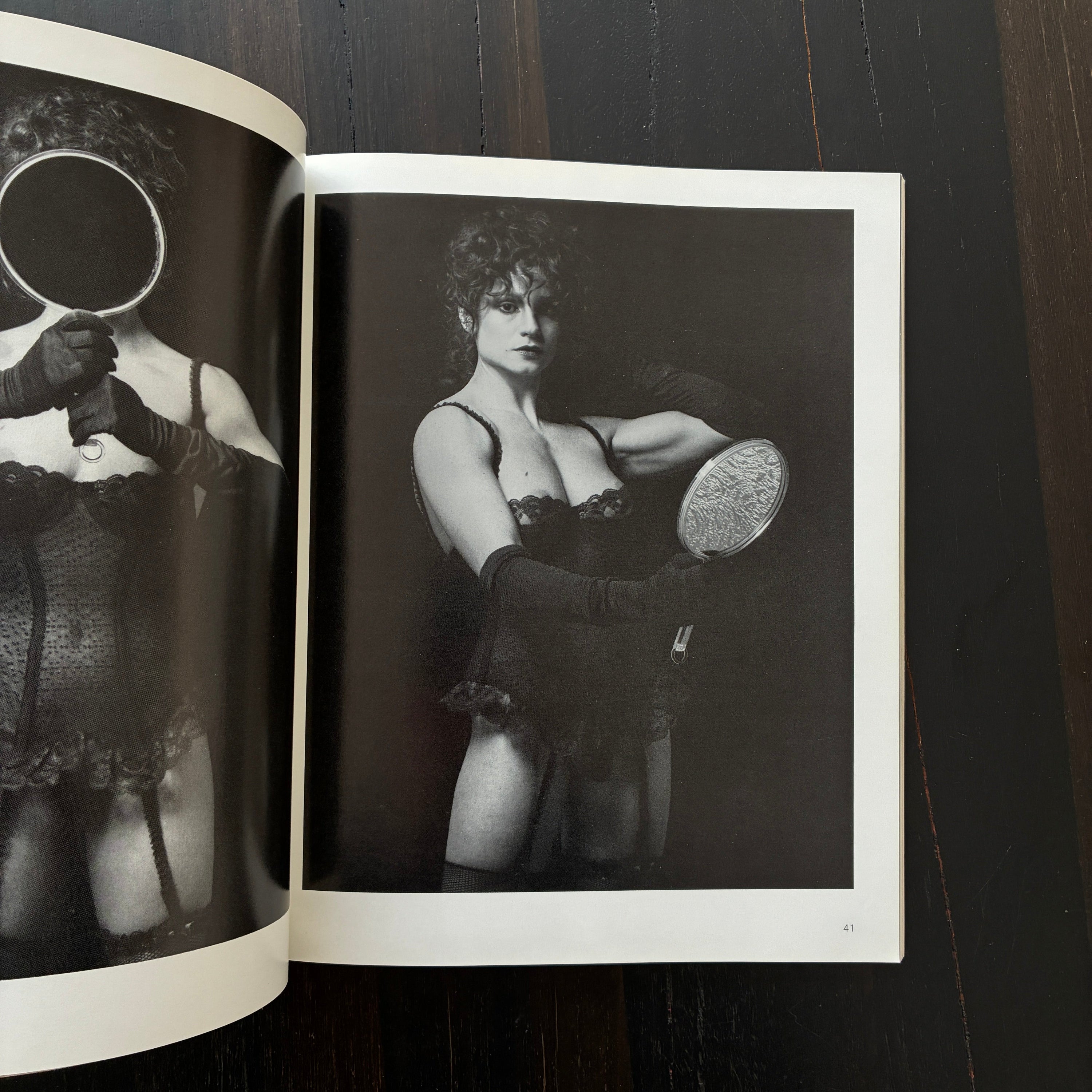 An open black-and-white art book or magazine laid flat on a dark wooden surface, showing glossy printed photographs of a woman in lingerie holding a mirror.